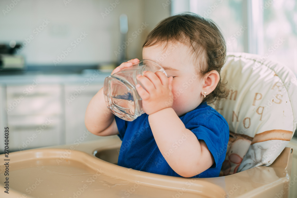 A little girl in the kitchen in the afternoon in a high chair drinking water from a glass glass A little girl in the kitchen in the afternoon in a high chair drinking water from a glass glass