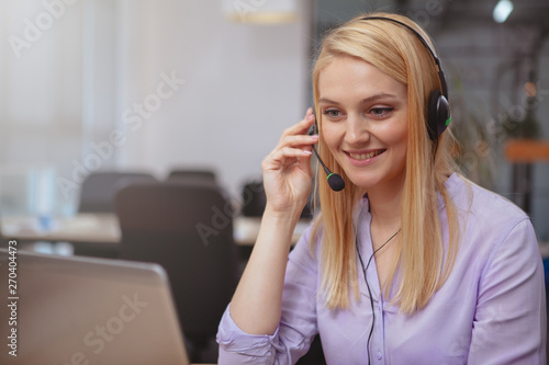 Fényképezés Beautiful cheerful woman smiling joyfully, answering customer calls at the call center, typing on her laptop
