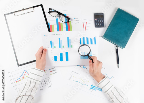 Woman's hands are studying the chart with a magnifying glass. Top view of business woman's hands on white desk with graphs. Finance and business concept.