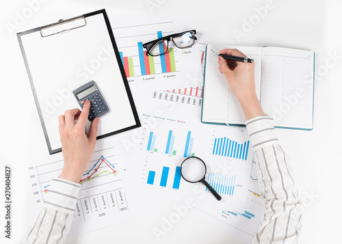 The hands of a woman count on a calculator and write notes. Top view of business woman's hands on white desk with graphs. Finance and business concept.