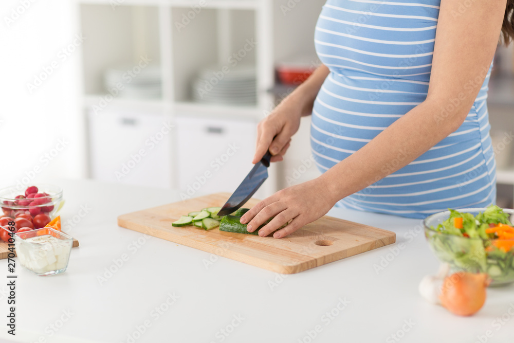 pregnancy, cooking food and healthy eating concept - close up of pregnant woman making vegetable salad and chopping cucumber by kitchen knife on cutting board at home kitchen