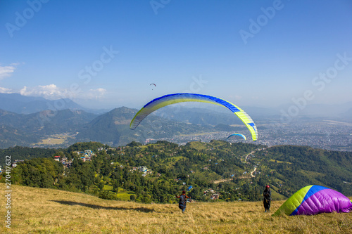 a paraglider takes off on a blue parachute from a hillside against the background of a city in a green mountain valley