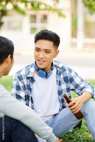 Young guy in casual clothing sitting on the grass with bottle of beer and talking to his friend outdoors