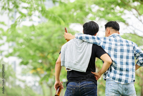 Rear view of two friends embracing each other and drinking beer while strolling in the park
