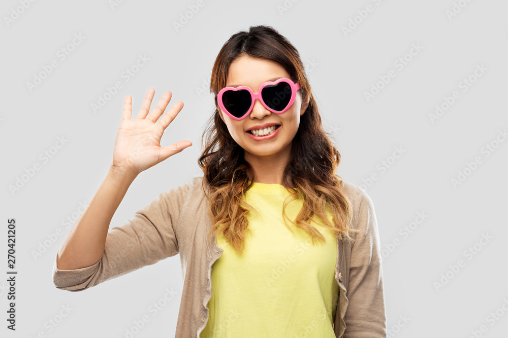 summer, valentine's day and eyewear concept - happy asian young woman in pink heart-shaped sunglasses waving hand over grey background summer, valentine's day and eyewear concept - happy asian young woman in pink heart-shaped sunglasses waving hand over grey background