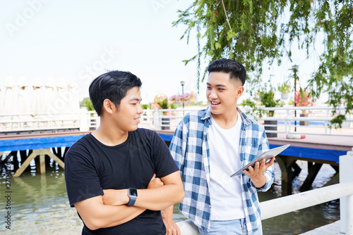 Two smiling teenagers using digital tablet and discussing something while standing on the pier outdoors