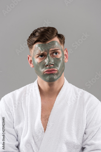 front view of sad young man in bathrobe with clay mask isolated on grey