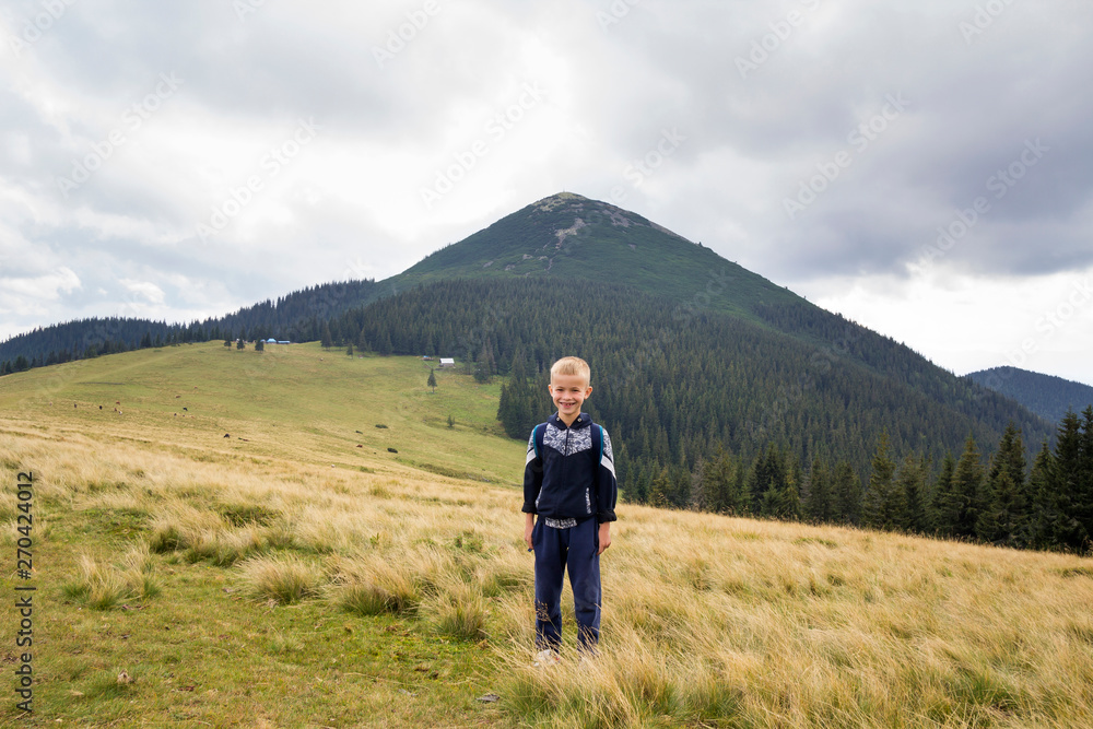 Young happy smiling child boy with backpack standing in mountain grassy valley on background of summer landscape, woody mountain view. Active lifestyle, adventure and weekend activity concept.