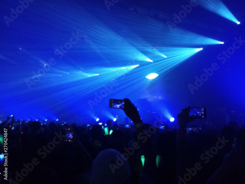 Blurred photo of people using a smart phone to take a photo of music festival and lights streaming down, rock concert
