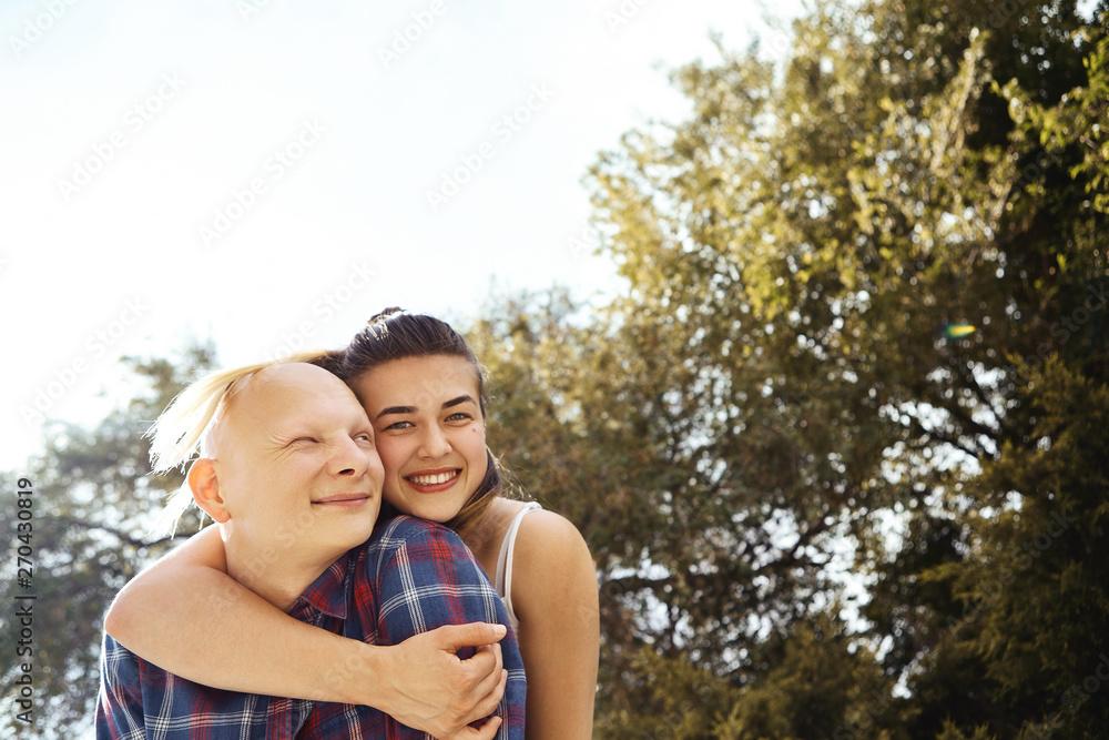 Portrait of young couple having fun and laughing in park. Girlfriend hugs her boyfriend from behind, smiling and looking straight at camera. Portrait of young couple having fun and laughing in park. Girlfriend hugs her boyfriend from behind, smiling and looking straight at camera.