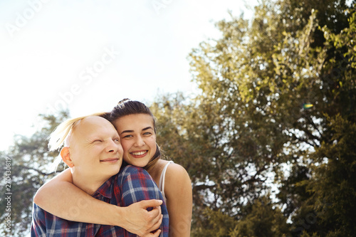 Portrait of young couple having fun and laughing in park. Girlfriend hugs her boyfriend from behind, smiling and looking straight at camera.
