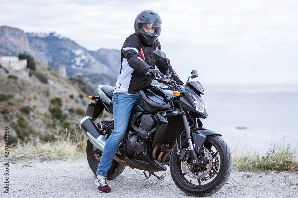 Young man biker with his black motorbike ready to drive, in front of the sea Young man biker with his black motorbike ready to drive, in front of the sea
