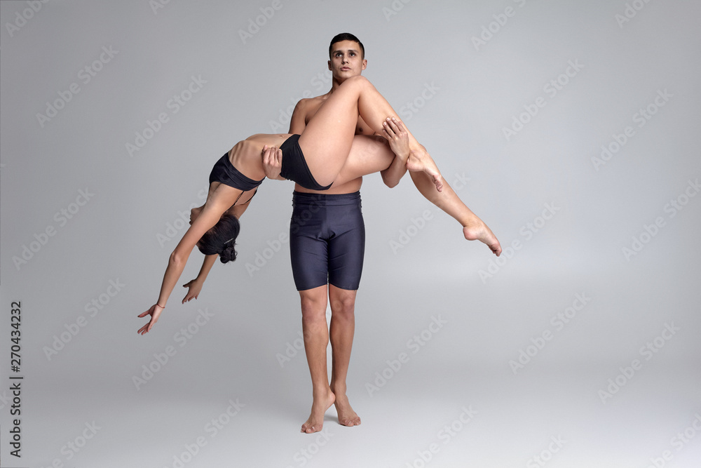 The couple of a young modern ballet dancers in black suits are posing over a gray studio background. The couple of a young modern ballet dancers in black suits are posing over a gray studio background.