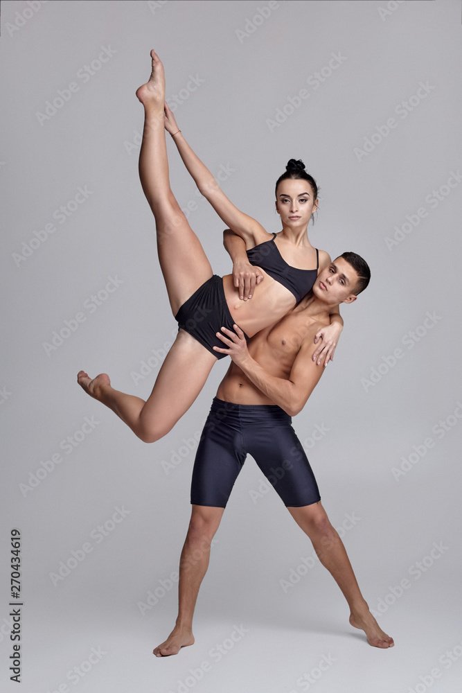 The couple of a young modern ballet dancers in black suits are posing over a gray studio background. The couple of a young modern ballet dancers in black suits are posing over a gray studio background.