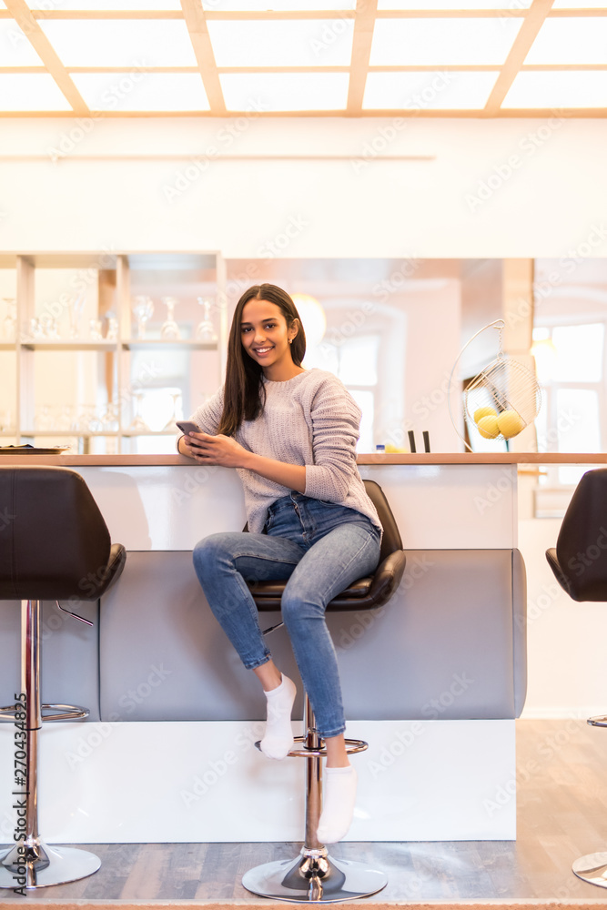 Young pretty woman using smartphone leaning at kitchen table in a modern home. Smiling woman reading phone message. Young happy girl typing a text message. Young pretty woman using smartphone leaning at kitchen table in a modern home. Smiling woman reading phone message. Young happy girl typing a text message.