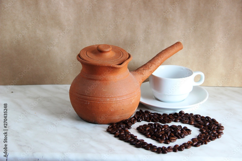 brown clay Turk for cooking Turkish coffee, white clean Cup and saucer and roasted coffee beans in the shape of a heart on the table
