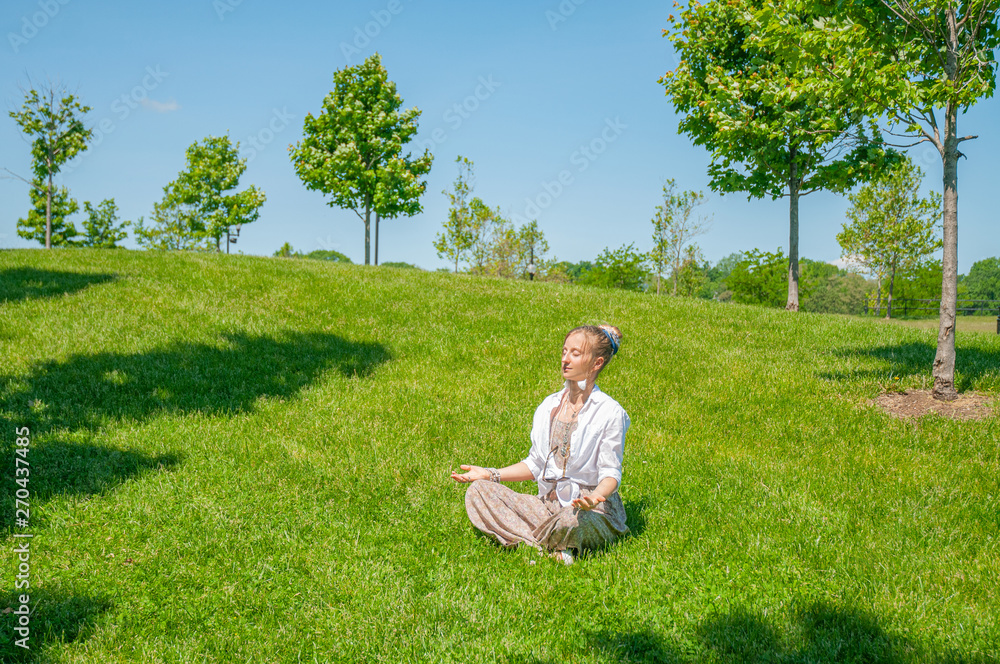 Beautiful boho style woman with accessories is meditating sitting in Lotus pose on grass in park. Beautiful boho style woman with accessories is meditating sitting in Lotus pose on grass in park.
