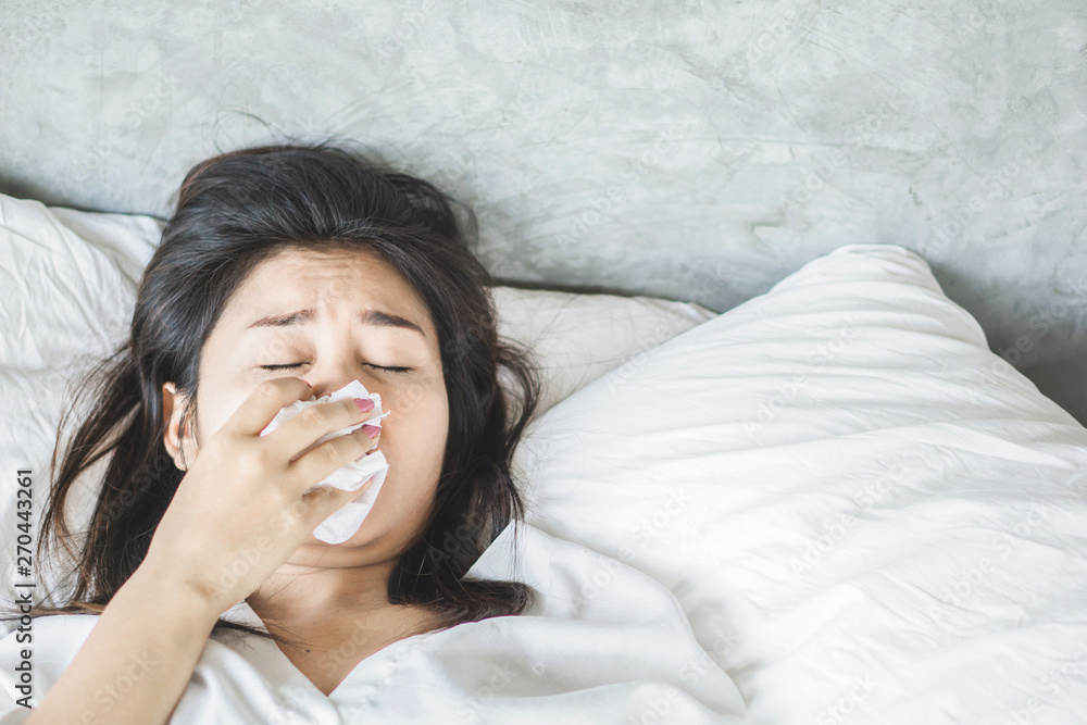 Asian woman having flu and sneezing feeling unwell lying down on bed with tissue in hand Asian woman having flu and sneezing feeling unwell lying down on bed with tissue in hand