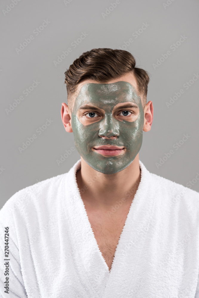 front view of smiling young man with clay mask in white bathrobe isolated on grey front view of smiling young man with clay mask in white bathrobe isolated on grey