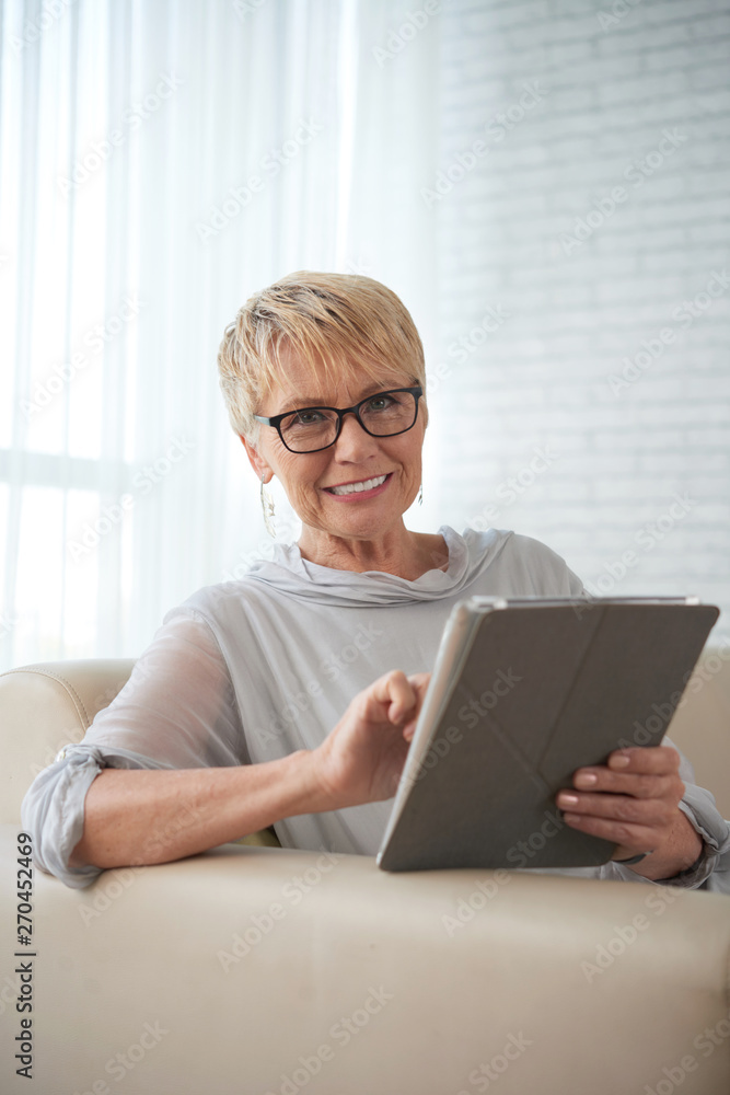 Portrait of mature woman with short hair wearing eyeglasses working online on digital tablet while sitting on sofa at home Portrait of mature woman with short hair wearing eyeglasses working online on digital tablet while sitting on sofa at home