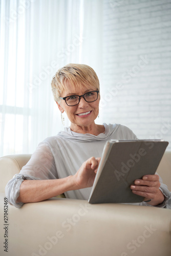 Portrait of mature woman with short hair wearing eyeglasses working online on digital tablet while sitting on sofa at home