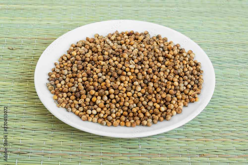 A lot of coriander seeds on a white saucer on a green table mat made of natural plant fibers. Natural food spices and seasonings. A lot of coriander seeds on a white saucer on a green table mat made of natural plant fibers. Natural food spices and seasonings.