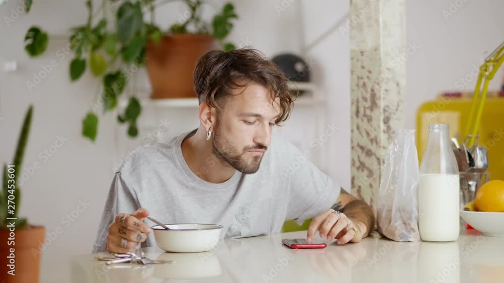 Nice tattooed millennial guy staying up to date in social media app while eating breakfast, pleasant man enjoying flakes with milk at morning time looking at news in smartphone display on table Nice tattooed millennial guy staying up to date in social media app while eating breakfast, pleasant man enjoying flakes with milk at morning time looking at news in smartphone display on table