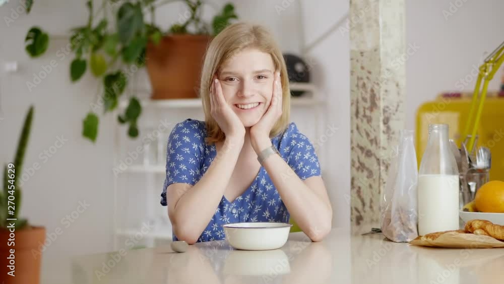 Portrait of thoughtful beautiful young woman dreaming in morning leaning on skinny arms, close up of female smiling looking to camera sitting in cozy light dining room with breakfast meal at table Portrait of thoughtful beautiful young woman dreaming in morning leaning on skinny arms, close up of female smiling looking to camera sitting in cozy light dining room with breakfast meal at table