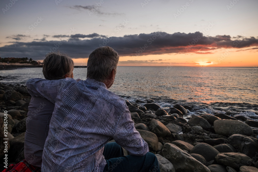Romantic elderly senior caucasian couple hug and enjoy the sunset at the beach - active mature retired people forever together stay in love with beautiful sky - happiness and vacation concept