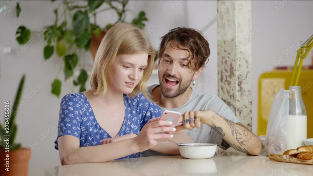 Couple of millennials discussing mail from friend using smart gadget at light dining room, beautiful girl touching blond hair and interesting guy talking explaining message text to lovely girlfriend Couple of millennials discussing mail from friend using smart gadget at light dining room, beautiful girl touching blond hair and interesting guy talking explaining message text to lovely girlfriend