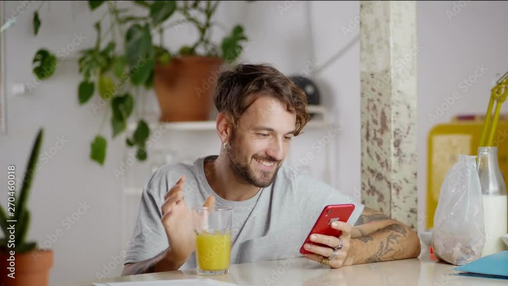 Nice Hispanic guy using new application at morning time while drinking juice from glass, green plant and column on background of casual male sitting at light kitchen busy with smart mobile device Nice Hispanic guy using new application at morning time while drinking juice from glass, green plant and column on background of casual male sitting at light kitchen busy with smart mobile device