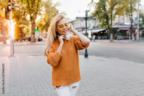 Outdoor portrait of stylish young woman in wool sweater listening music with eyes closed and smile. Wonderful girl in white ripped pants walking down the street in good mood.