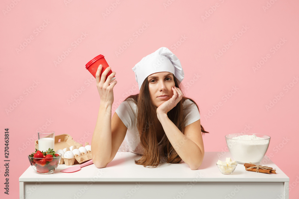 Sad tired chef cook confectioner or baker in apron white t-shirt, toque chefs hat sit at table with paper cup of coffee isolated on pink pastel background in studio. Mock up copy space food concept. Sad tired chef cook confectioner or baker in apron white t-shirt, toque chefs hat sit at table with paper cup of coffee isolated on pink pastel background in studio. Mock up copy space food concept.