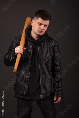 The young man wearing leather jacket standing in fighting pose with baseball bat against dark background in studio