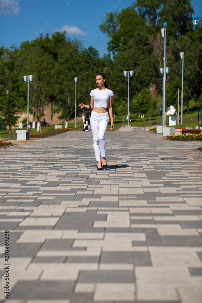 Young beautiful brunette girl in white jeans and t-shirt walking in summer park