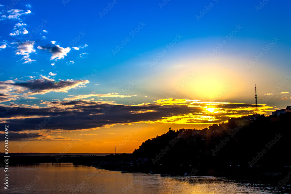 Silhouette of monument to Salawat Yulaev against a bright yellow blue sky with clouds and orange sun disk at city sunset on the river bank. Salavat Yulayev, Ufa, Bashkortostan, Russia.