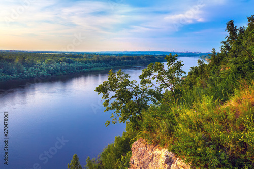 A large cliff overgrown with green grass and trees dominate over the White River in the evening at dusk during sunset under a clear blue sky. Rock Hanging Stone, Ufa, Bashkortostan, Russia.