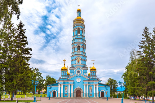 Tableau sur toile Light blue high church with golden domes surrounded by green trees under large rain clouds on the square