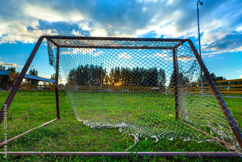 A small football goal with a torn net on green grass against a blue sky with white clouds and a yellow sun hiding behind the trees. Biathlon Stadium, Ufa, Bashkortostan, Russia.