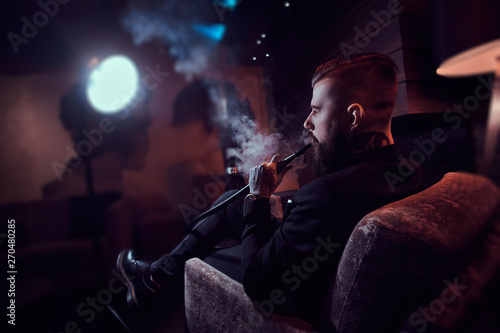 Brutal tattooed man is making nice misty vapour while relaxing near fireplace and smoking hookah.