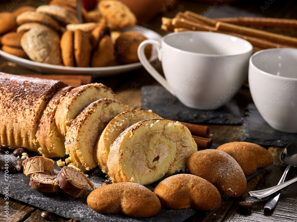 Fototapeta premium Sand cookies heart shape, rolled cake with cherry and cinnamon stick, coffee cups on kitchen on wooden table in village style. Useful for digestion. Limitations for diabetics.