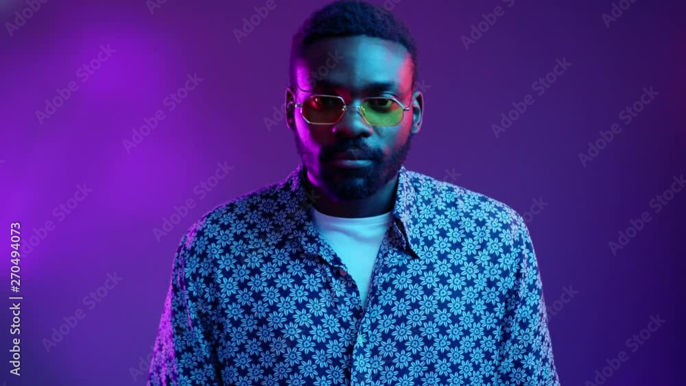 Serious African American young man with yellow glasses look at camera posing on the blue and violet neon light background attractive beard black blue boy bright cheerful close up slow motion Serious African American young man with yellow glasses look at camera posing on the blue and violet neon light background attractive beard black blue boy bright cheerful close up slow motion