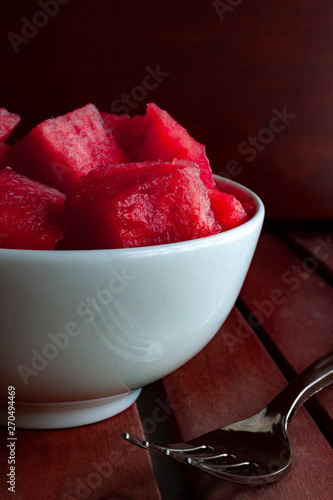 WATERMELON IN PIECES IN WHITE BOWL WITH FORK ON WOODEN TABLE