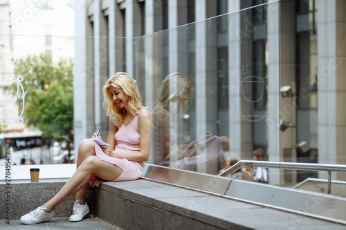 Happy young blonde woman in rose dress and white sneakers sit on the floor and write down something in notebook opposite building in city center
