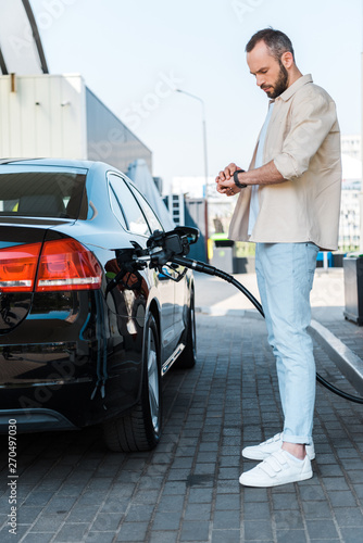 Handsome man standing and looking at watch while refueling black car at gas station