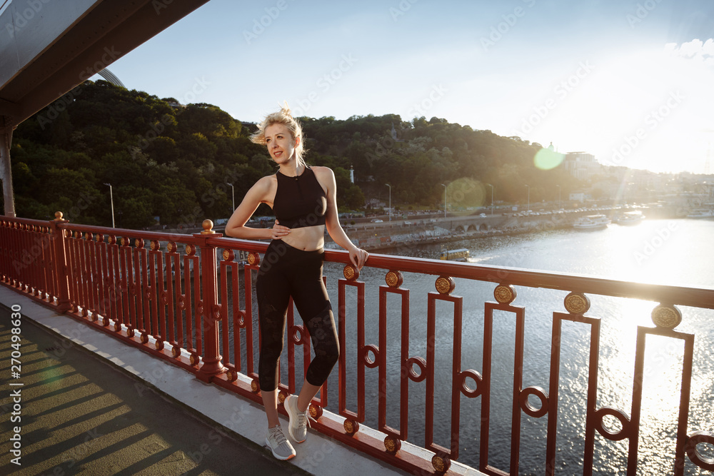 Beautiful happy blonde woman in black sportswear walks on the bridge after fitness training above the river at sunset.