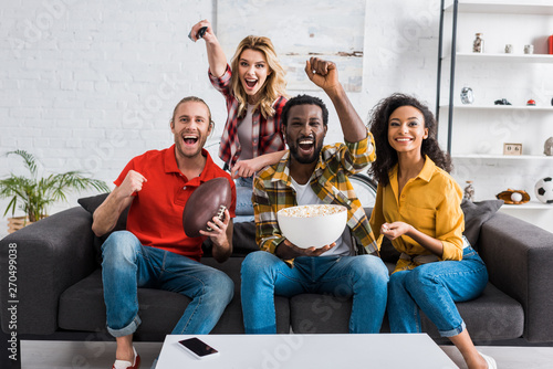 Happy multiethnic friends sitting on couch, watching match and holding bowl of popcorn