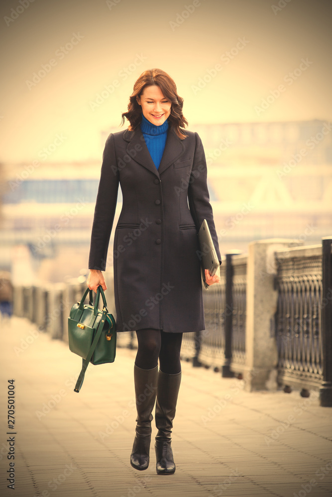 A beautiful smiling middle-aged woman in a black coat with a laptop and a green hand bag