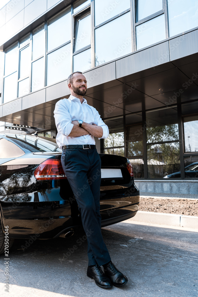 low angle view of handsome man standing with crossed arms near black car low angle view of handsome man standing with crossed arms near black car