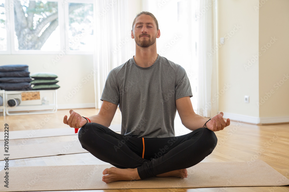 Man meditating and holding hands in mudra gesture in class. Handsome young guy practicing yoga and sitting with his legs crossed. Meditation and yoga class concept. Front view. Man meditating and holding hands in mudra gesture in class. Handsome young guy practicing yoga and sitting with his legs crossed. Meditation and yoga class concept. Front view.